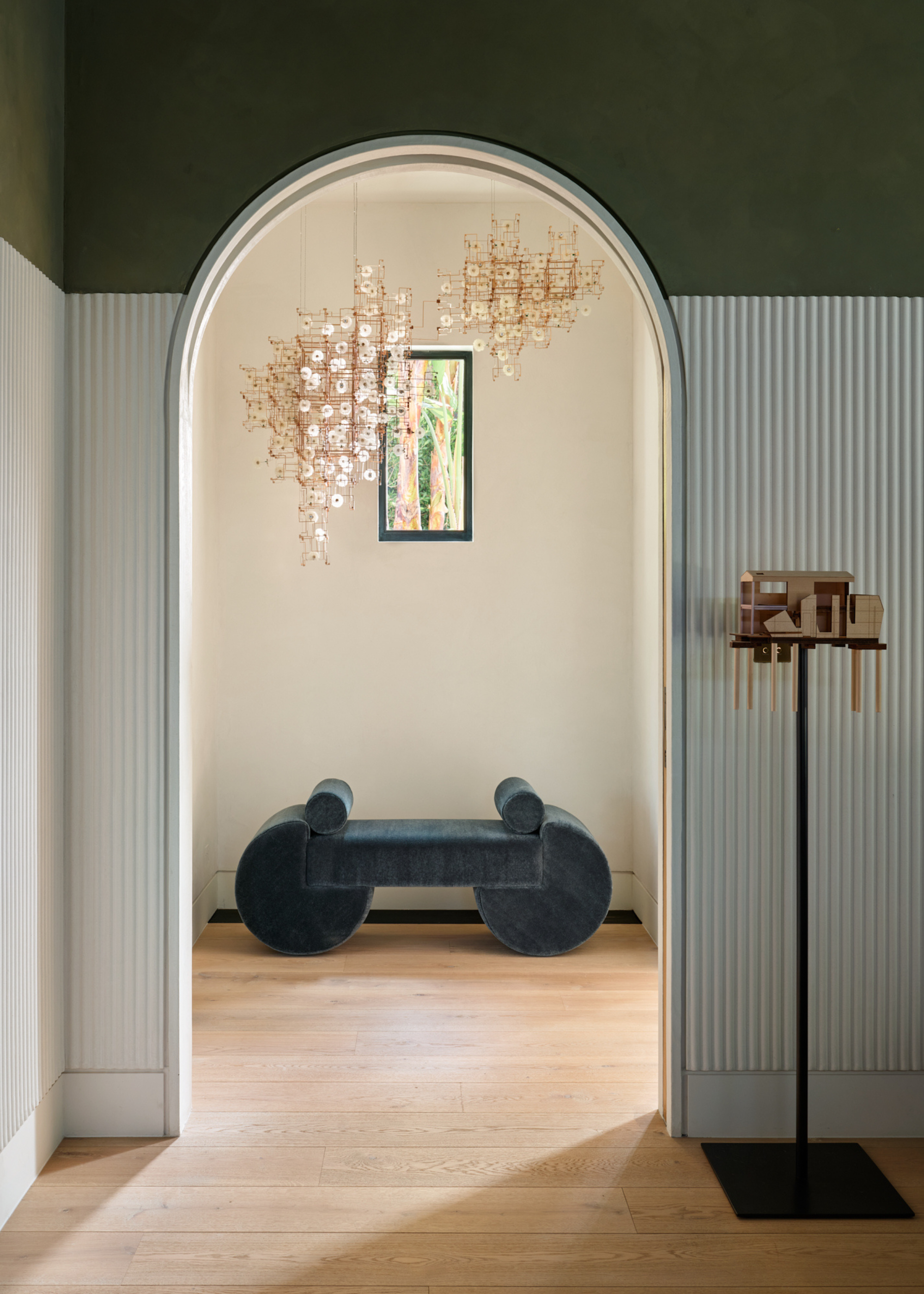 A room with an arched doorway featuring a deep green and white ribbed wall leading to a room with a blue suede bench and hanging chandeliers