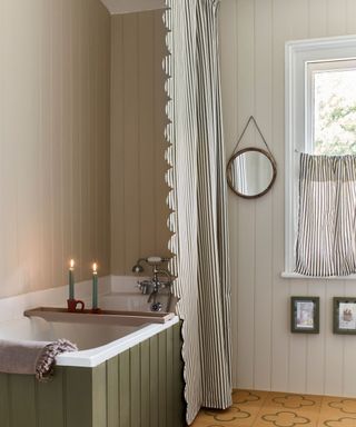 A bathroom with greige shiplap paneling, a scalloped striped curtain and blind, and an olive green paneled bath tub with a white interior and a bath tray with two blue lit candles on it