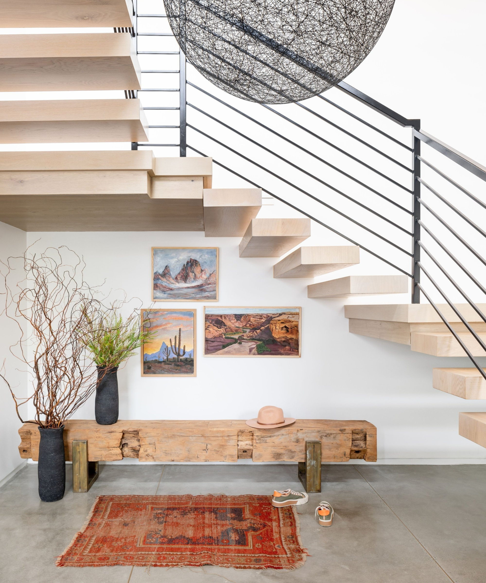 an entryway sitting area underneath a set of stairs that features a distressed wooden bench, a small distressed persian rug, two vases holding plant life, and some artwork hanging on the wall
