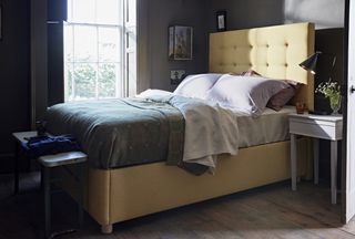 small bedroom with dark gray walls, yellow upholstered bed in corner by window, shutters, bare floorboards, small bench, nightstand, blanket, taxidermy on wall