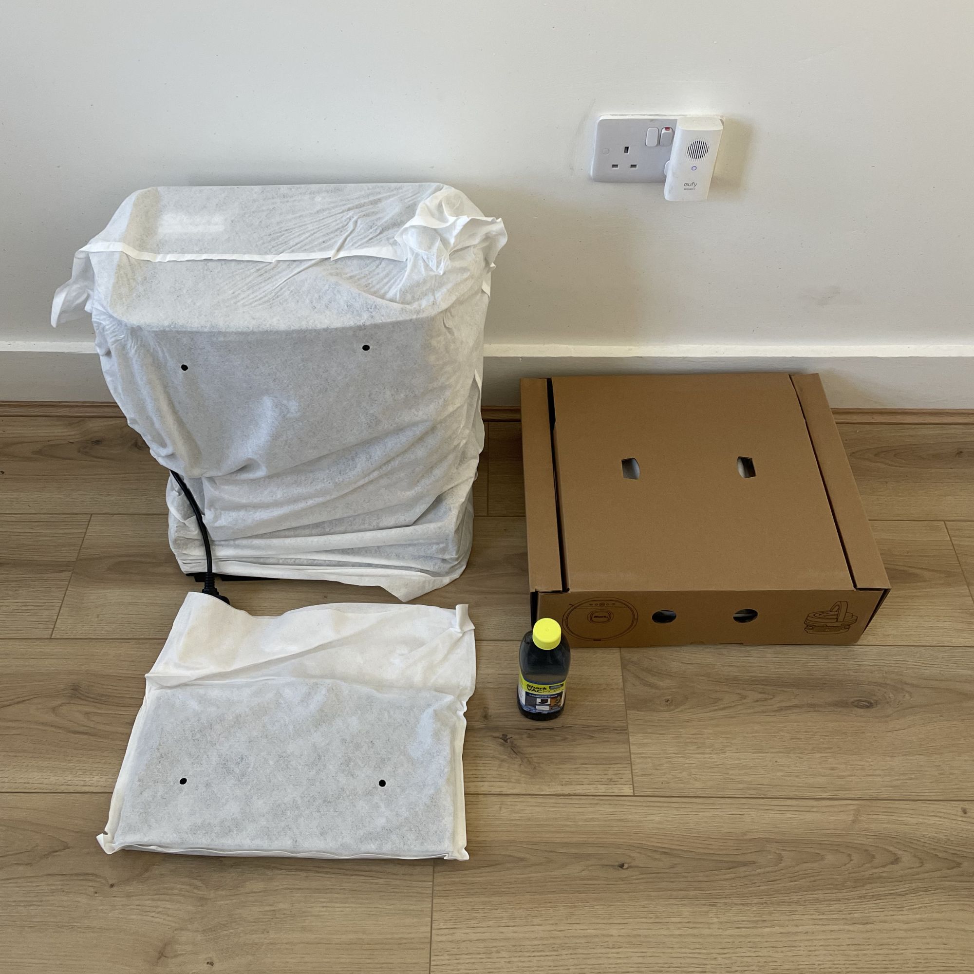Components of the Shark PowerDetect UV Reveal 2-in-1 Vac & Mop Robot Vacuum sitting on the floor, wrapped in packaging