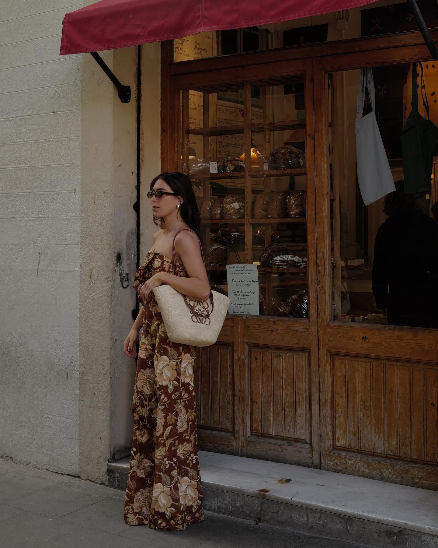 Woman wears floral print matching top and trousers, carries woven raffia basket bag by Loewe