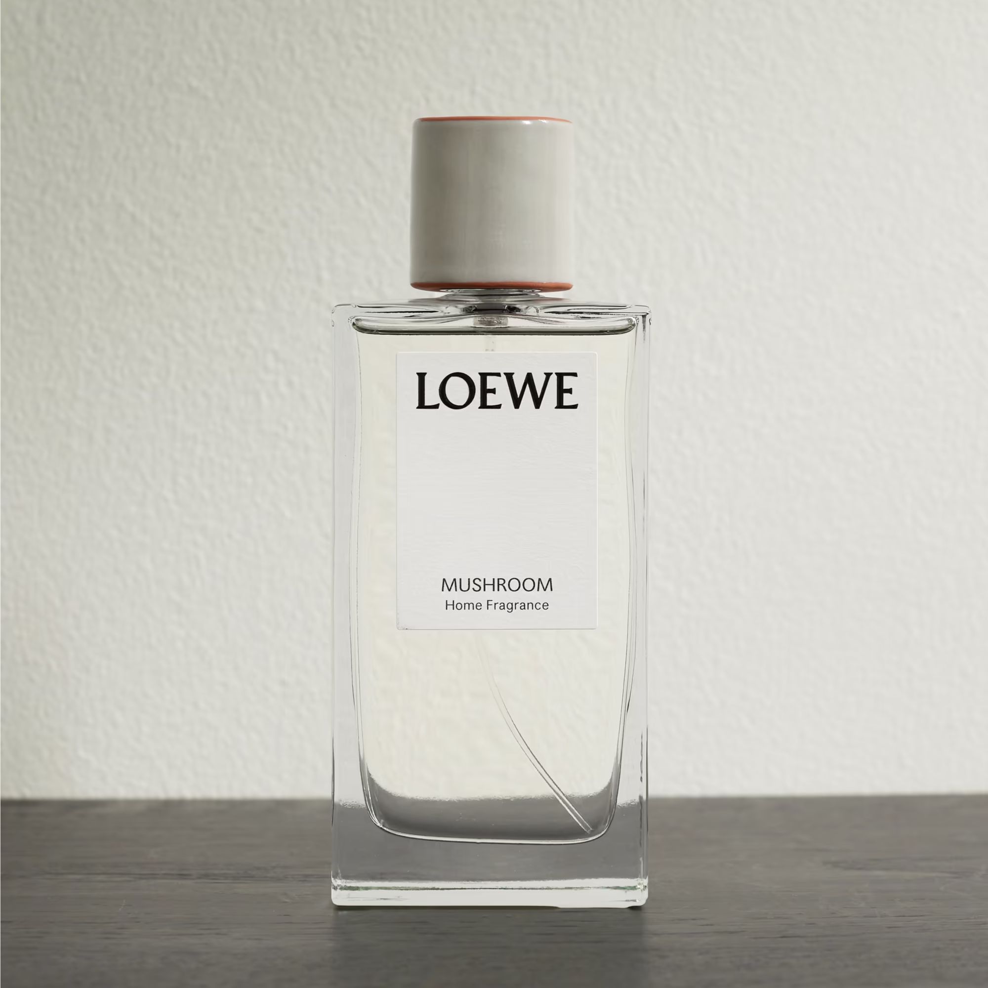 A glass bottle of Loewe Mushroom room spray against a white wall on a wooden surface
