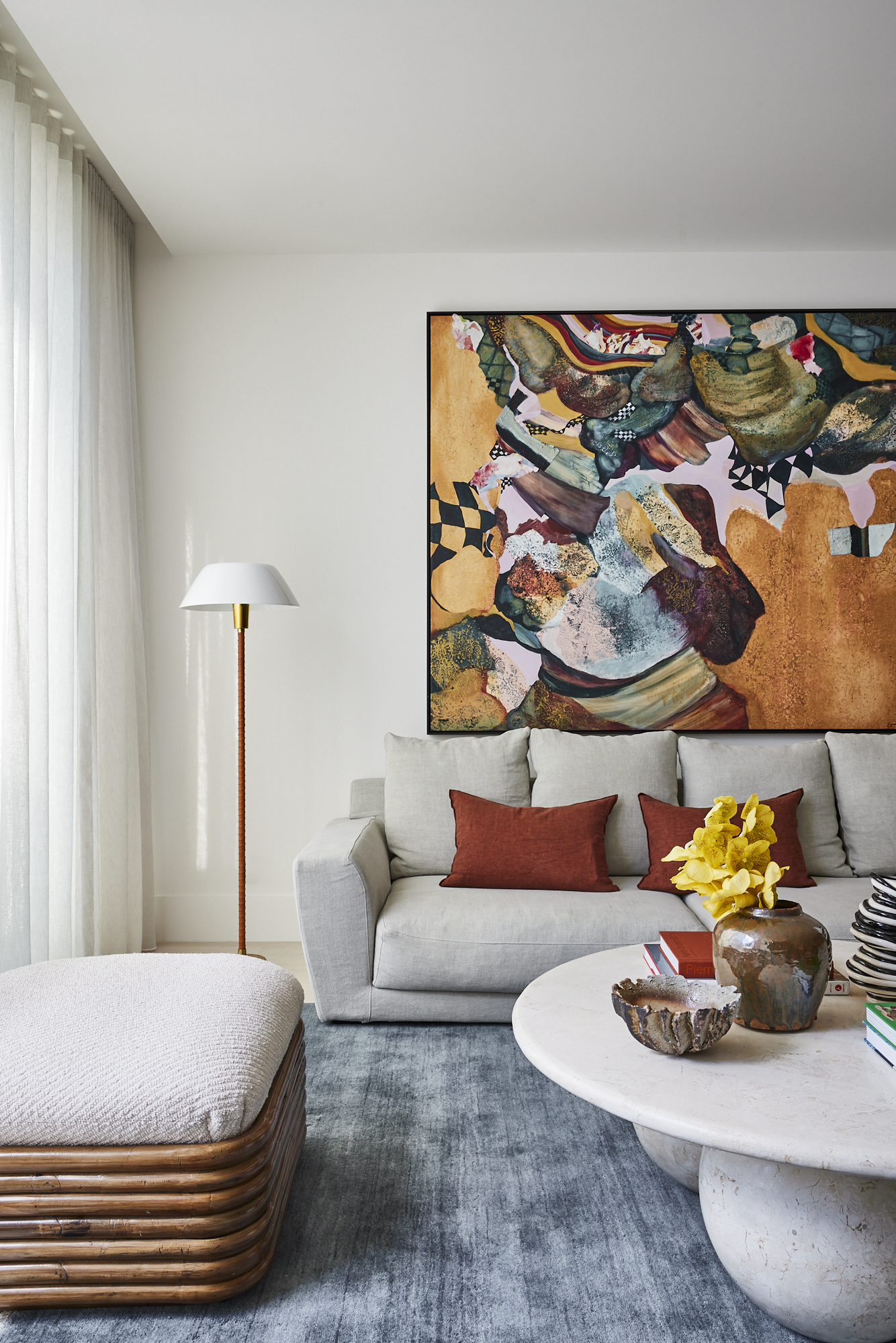 beige living room with blue rug, colorful artwork, beige sofa with orange cushions, white coffee table styled with vases and books, and a beige ottoman with rattan details