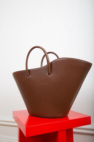 Brown open tote bag inspired by the structure of an oyster shell, placed on a red geometric shaped stool against a cream background