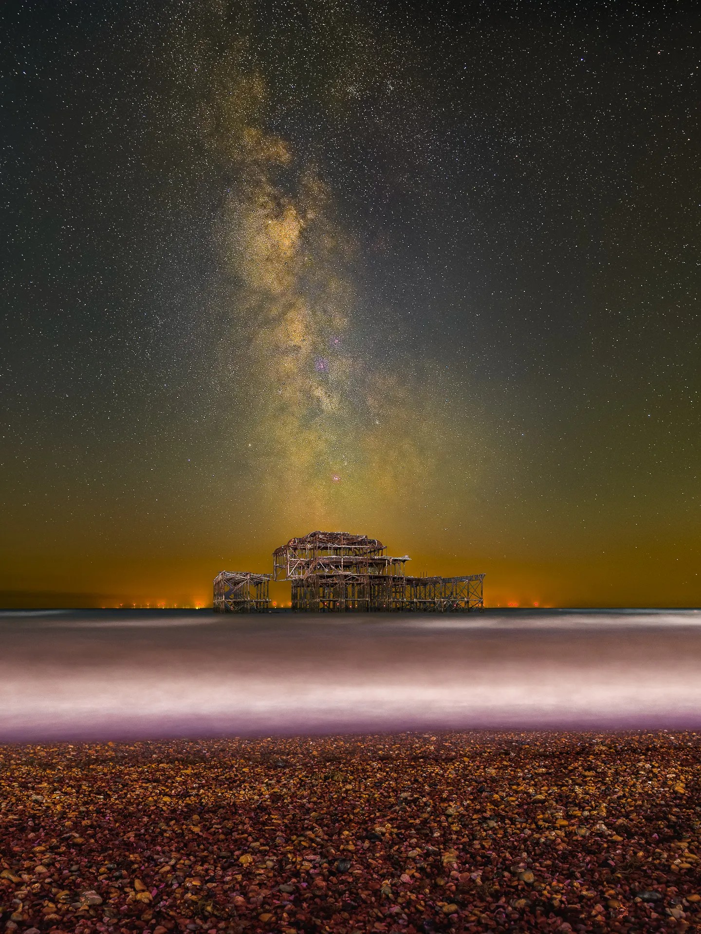 Night photograph of the Milky Way galaxy arcing above the decaying structure of the West Pier in Brighton, with the sea blurred from a long exposure and a pebble beach in the foreground.