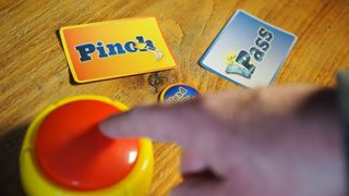 A hand touches a buzzer laid out on a wooden table, with cards reading 'pinch' and 'pass' visible in the background