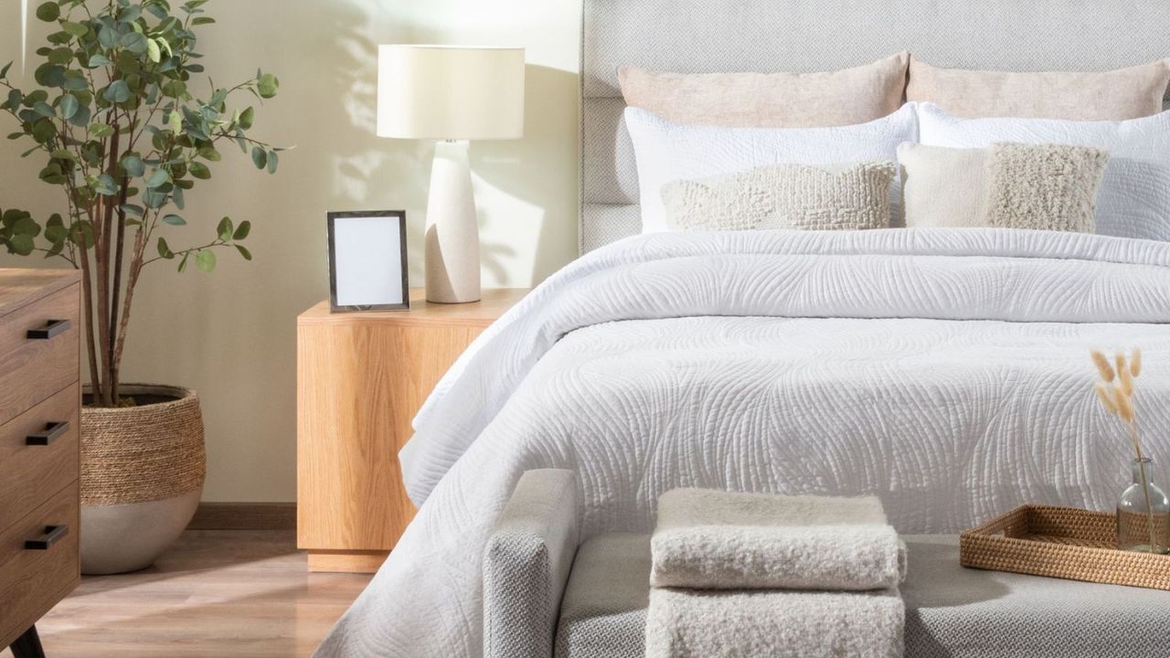 A bright bedroom with grey bed, storage ottoman with wooden tray and beige blanket and carpet, with wooden chest of drawers and bedside table, tall houseplant, white lamp, empty picture frame, off-white bedding, cushions and abstract wall art