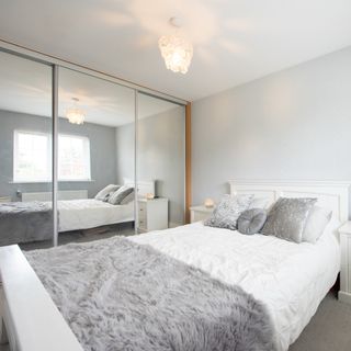 A general interior view of a bedroom painted grey, with a white wooden bed frame, fur throw blanket, crushed velvet square and round cushions, bedside unit with candle, mirrored sliding door wardrobe, window with venetian blinds within a home