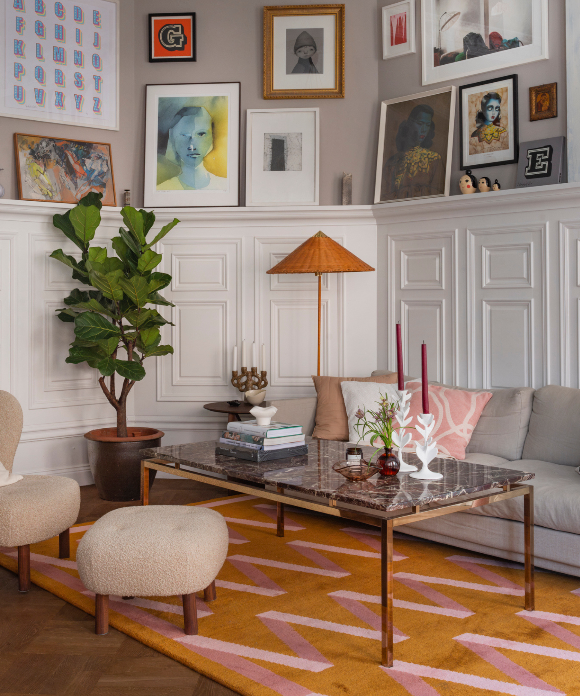 a living room with lot of artwork hung on the walls, a large coffee table with a marble top, a grey couch, an accent chair, and an orange and pink rug
