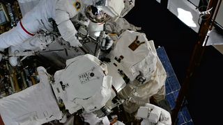 Two astronauts in white spacesuits work to repair a robotic arm outside of a space station
