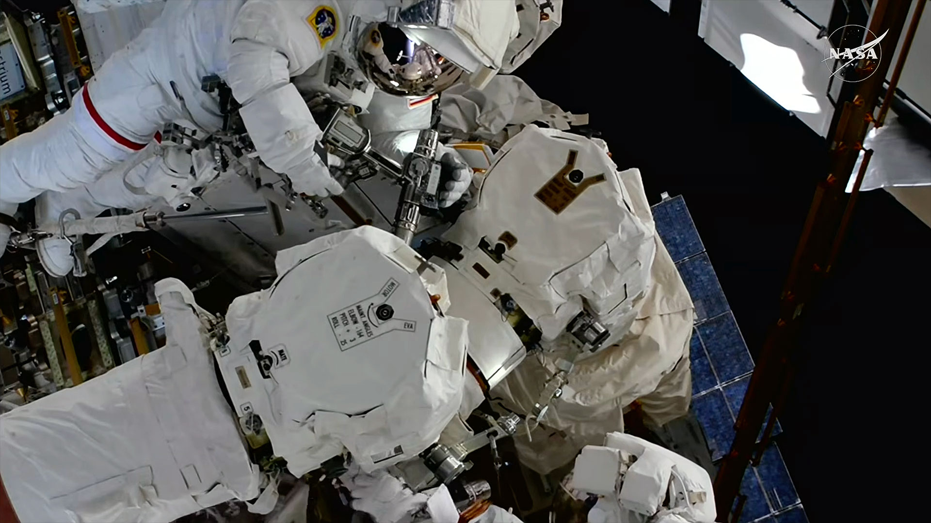 Two astronauts in white spacesuits work to repair a robotic arm outside of a space station