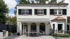 exterior of white house with black-and-white striped awning over outdoor dining area, with arched windows, an outdoor kitchen, pavers, and black fence