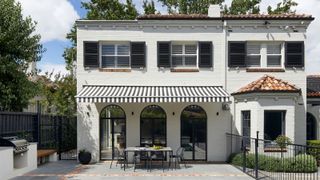 exterior of white house with black-and-white striped awning over outdoor dining area, with arched windows, an outdoor kitchen, pavers, and black fence
