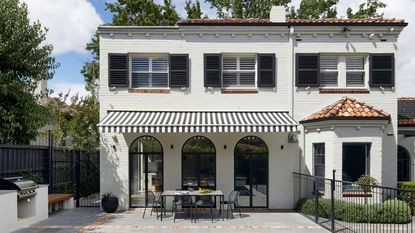 exterior of white house with black-and-white striped awning over outdoor dining area, with arched windows, an outdoor kitchen, pavers, and black fence