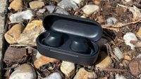 A black pair of Sony WF-C510 in-ear headphones inside their charging case with the lid open. They are positioned on a surface made up mainly of stones.