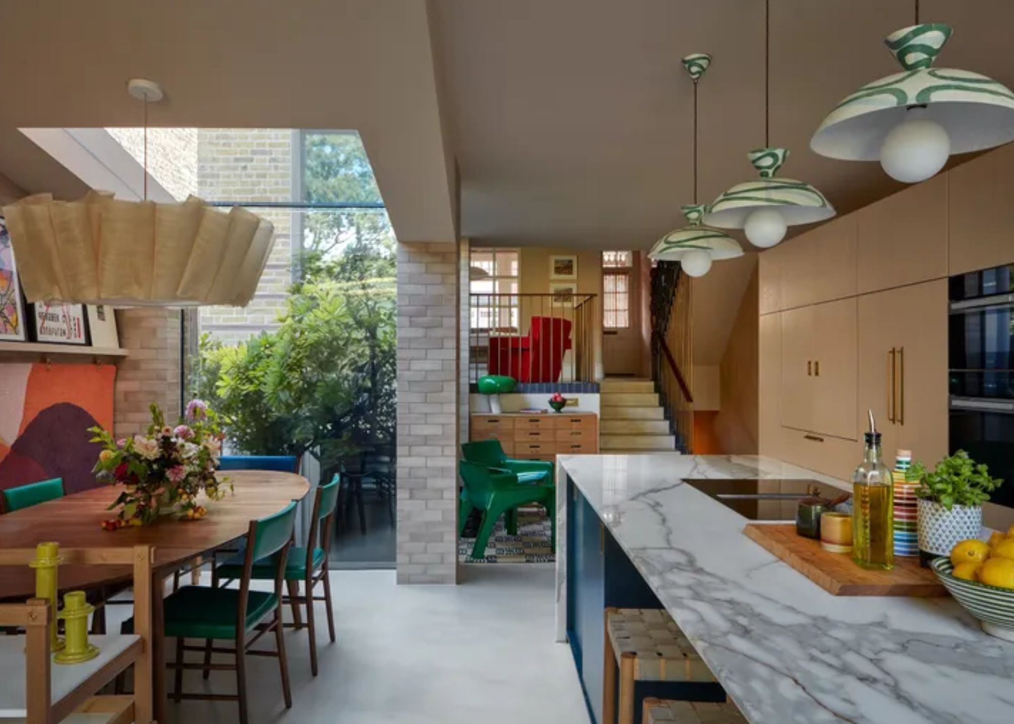 A marble island and timber dining table. Above the island are green and white shaded light pendants. Above the table is a large statement pendant.