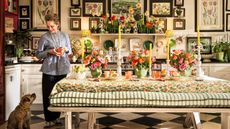 Butter Wakefield smiles in her maximalist kitchen, holding a drink next to a long dining table set with colorful floral arrangements, yellow candles, and a patterned tablecloth. A small terrier sits at her feet