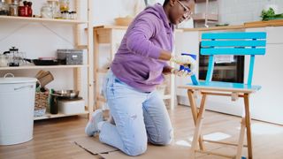 Woman kneeling on folding cardboard mat in kitchen while painting an outdoor chair with paintbrush