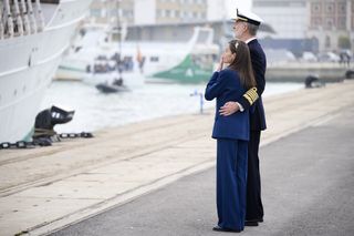 Queen Letizia wearing a blue trouser suit covering her mouth with her hand standing with King Felipe who has his arm around her as they look at a ship on the water