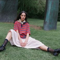 A woman sat on the grass wearing a long polka dot skirt, red and blue striped t-shirt, and beige neckerchief from Damson Madder.