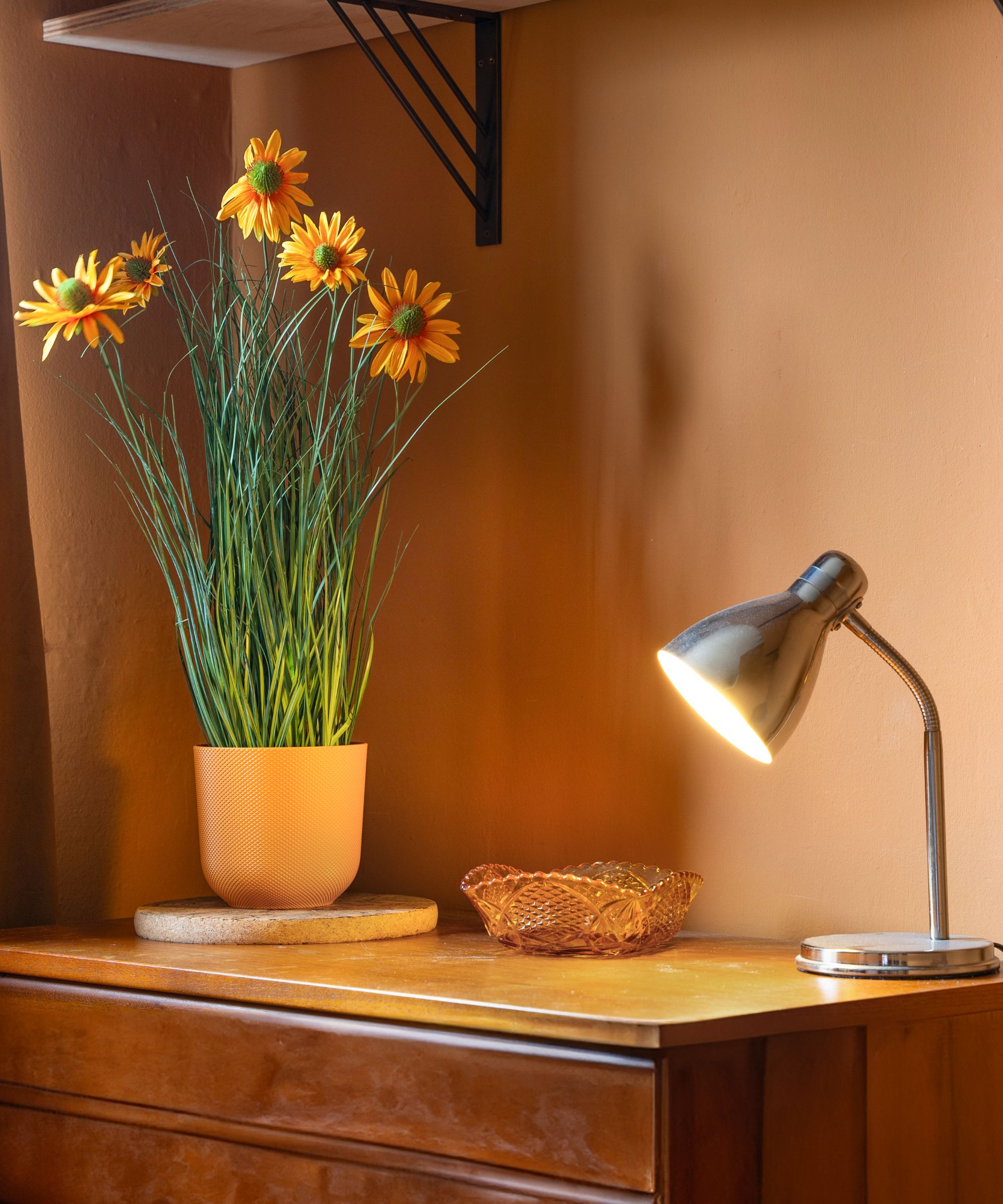 A tabletop lamp is lit on top of a chest of drawers, next to a vase of artificial sunflowers and a small item tray, against brown walls.