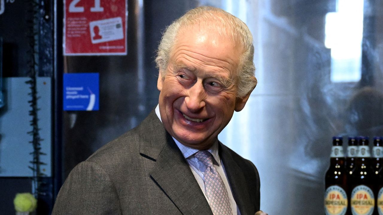 King Charles wearing a gray suit and smiling at a bar 