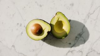 An avocado cut in half on top of a white marble kitchen worktop