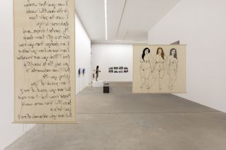A gallery area with two hanging canvases. Left: A scroll-like piece with written Arabic text. Right: Three naked woman drawn standing next to each other.