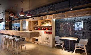 Odessa, Kiev, Ukraine. A restaurant with a wooden counter with high chairs, a dining table and chairs, wooden wall shelving with produce and glasses on it and pendant lights.