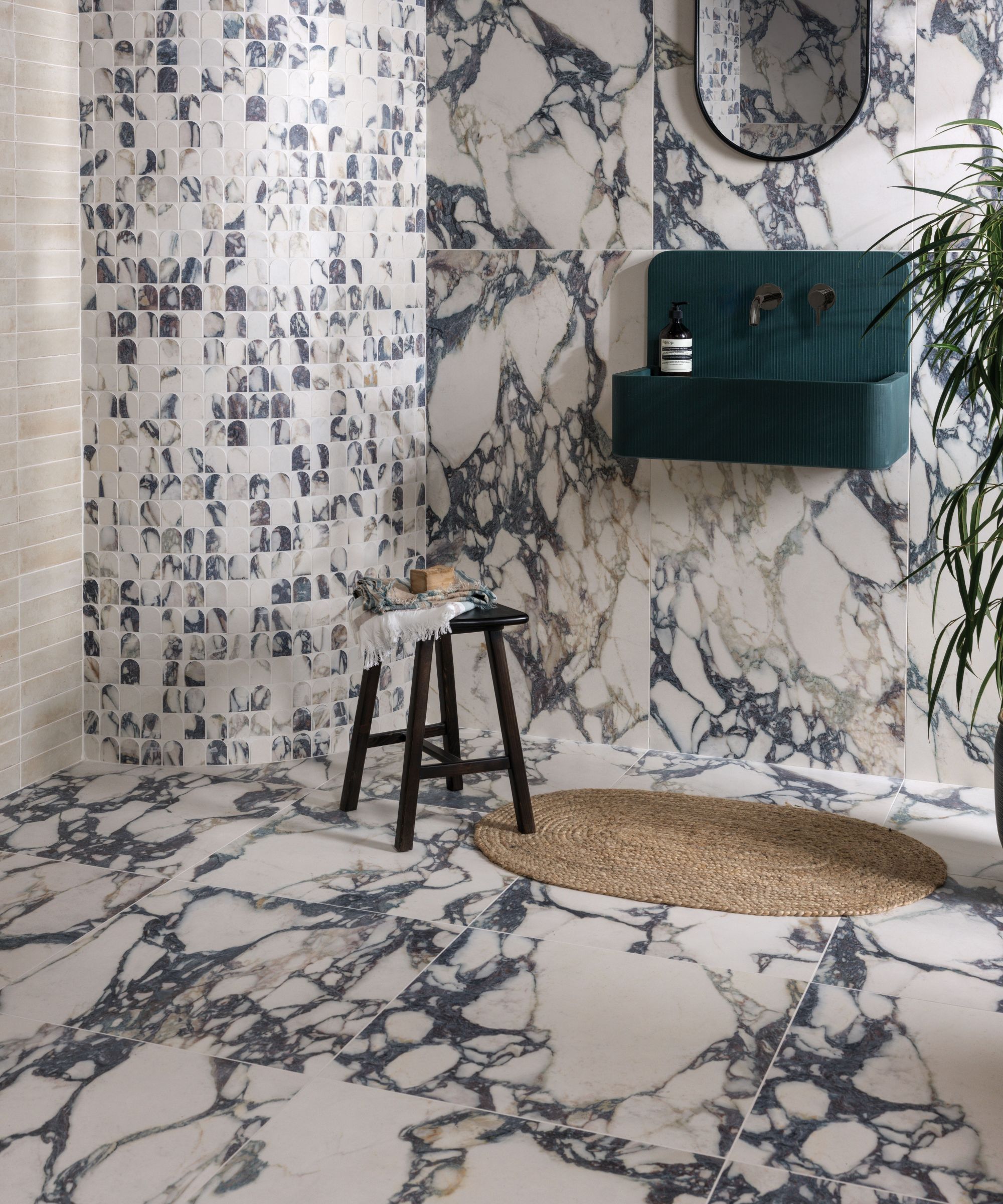 A dramatic bathroom combining large-format white and navy Calacatta marble tiles on floors and walls with a feature wall of small arch-shaped mosaic tiles in the same marble. A deep teal fluted wall-mounted basin, black oval mirror, dark wood stool and woven jute rug complete the bold, luxurious scheme.