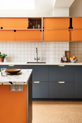 Kitchen with dark navy lower cabinets, orange upper cabinets, white square backsplash tiles and cork floor