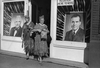 Black and white image, Greenfield, Indiana. 1960, two mature smiling ladies finely dressed coming out of voting station, two male candidate images in the shop windows