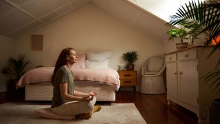 A woman sits at the foot of her bed practising a relaxing deep breathing technique to help her fall asleep