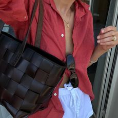 fashion influencer Lucy Williams takes an outfit selfie with a delicate gold necklace, red linen button-down shirt, black sunglasses, black woven Dragon Diffusion tote bag, and white cotton shorts