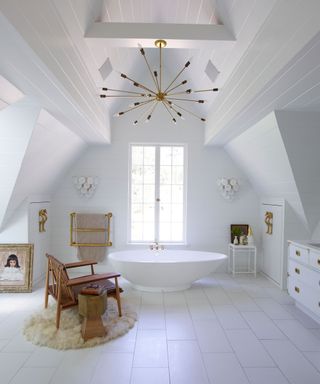 A large with bathroom with a white bath below a window, a wooden armchair and white floor tiles