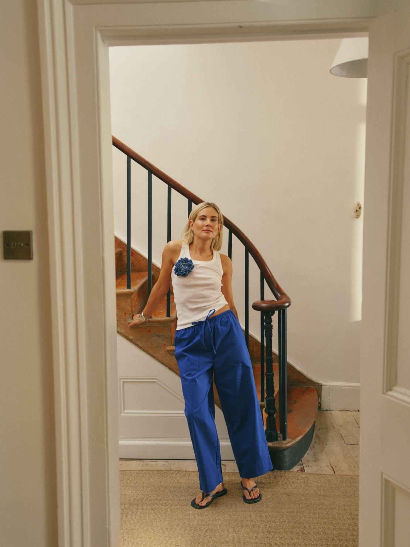 Influencer @lucywilliams02 poses by a staircase wearing cobalt blue trousers with a white vest top and blue flip flops.