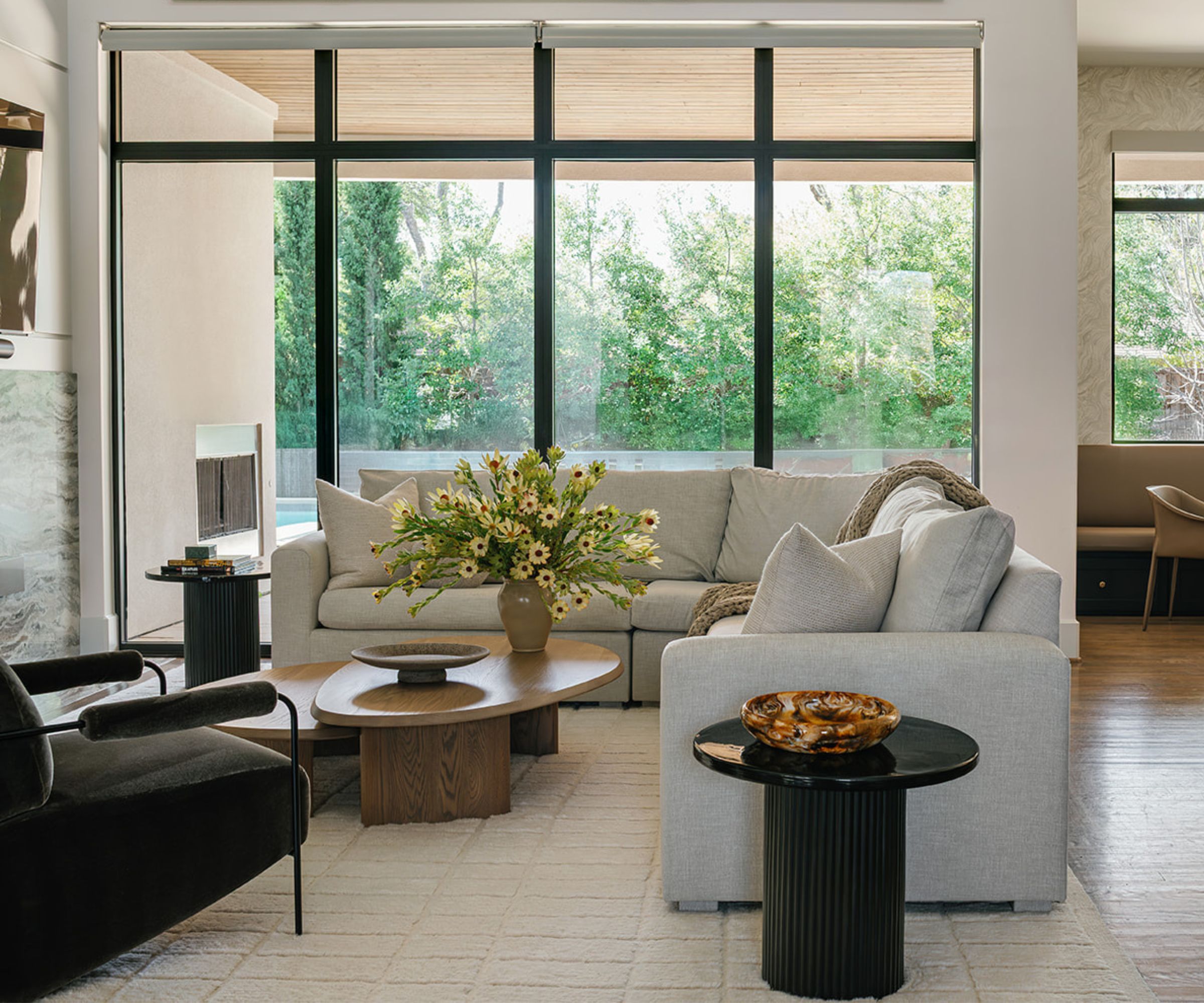 warm minimalist neutral open plan living room with large glass windows, neutral rug with a neutral L shaped section, wooden rounded coffee table and round black side table