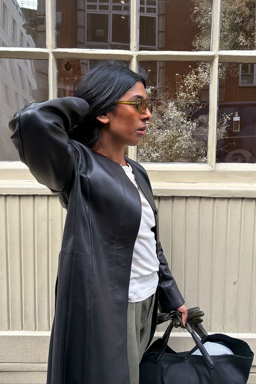 A woman standing outside in front of a storefront wearing a black leather duster jacket over a white T-shirt, green cargo pants, a black tote, and green oval sunglasses.