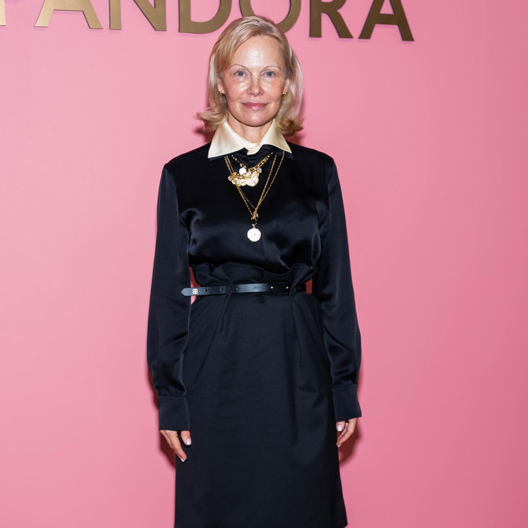 Pamela Anderson standing in front of a pink wall wearing a black long-sleeved top with a champagne color peaking through the top. 