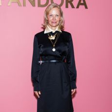 Pamela Anderson standing in front of a pink wall wearing a black long-sleeved top with a champagne color peaking through the top.