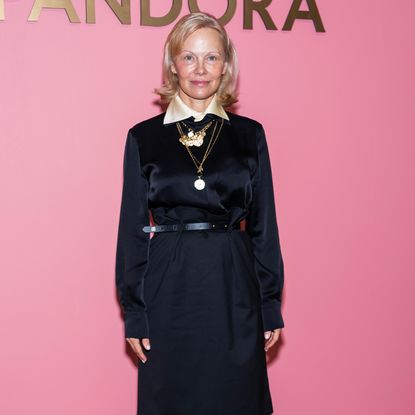 Pamela Anderson standing in front of a pink wall wearing a black long-sleeved top with a champagne color peaking through the top. 