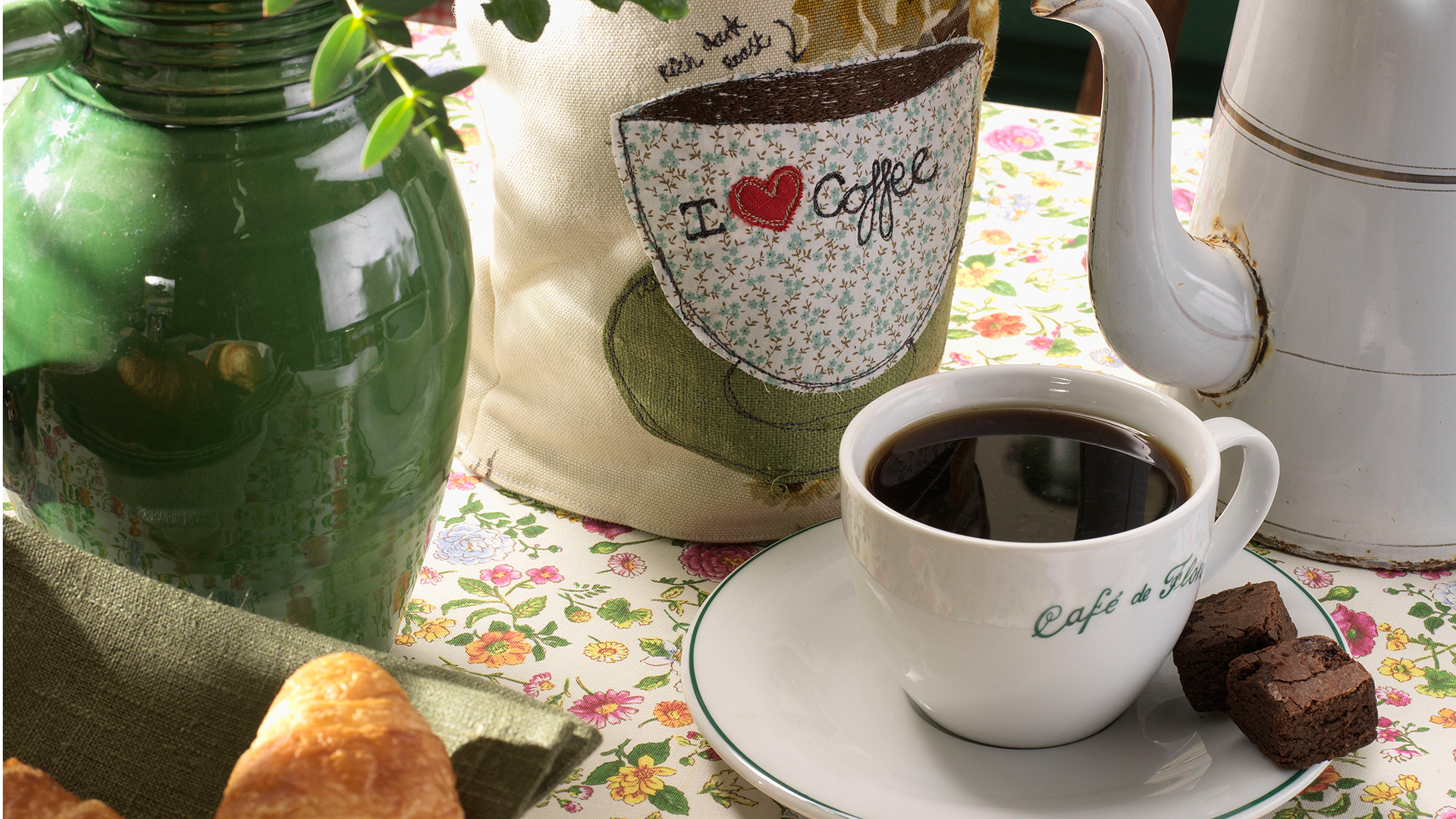 Garden table with a floral tablecloth with a cup of coffee with cafe written on it and a coffee pot cosy that says I love coffee next to it