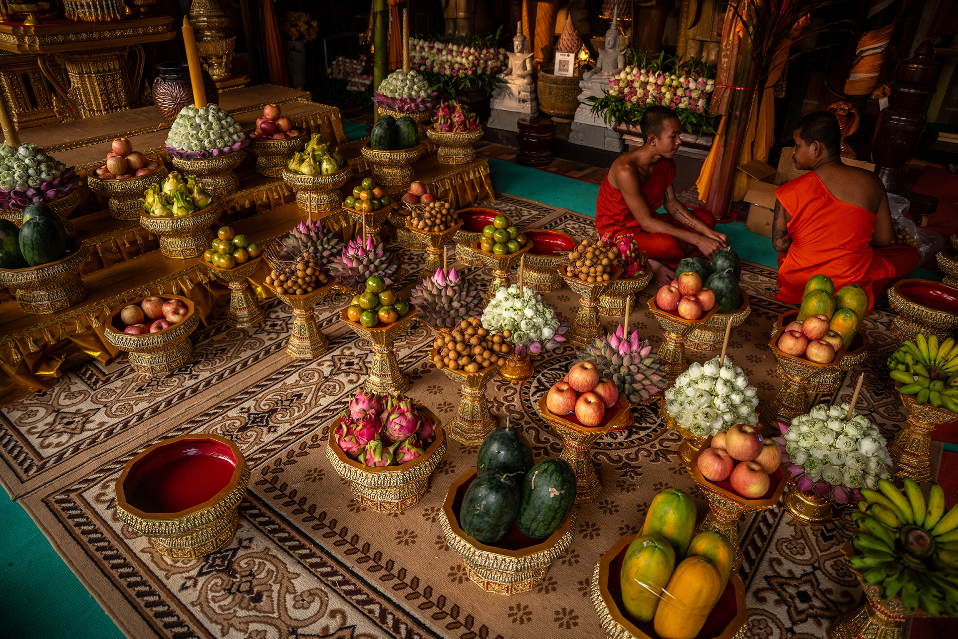 A vibrant display of fruits, flowers, and decorative bowls arranged on a patterned rug in a temple setting, reflecting cultural traditions