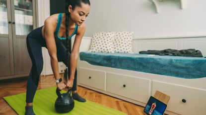 woman standing on a green exercise mat in a home setting holding a kettlebell that's on the floor in between her lets. she's looking at an ipad in front on the floor in front of her.