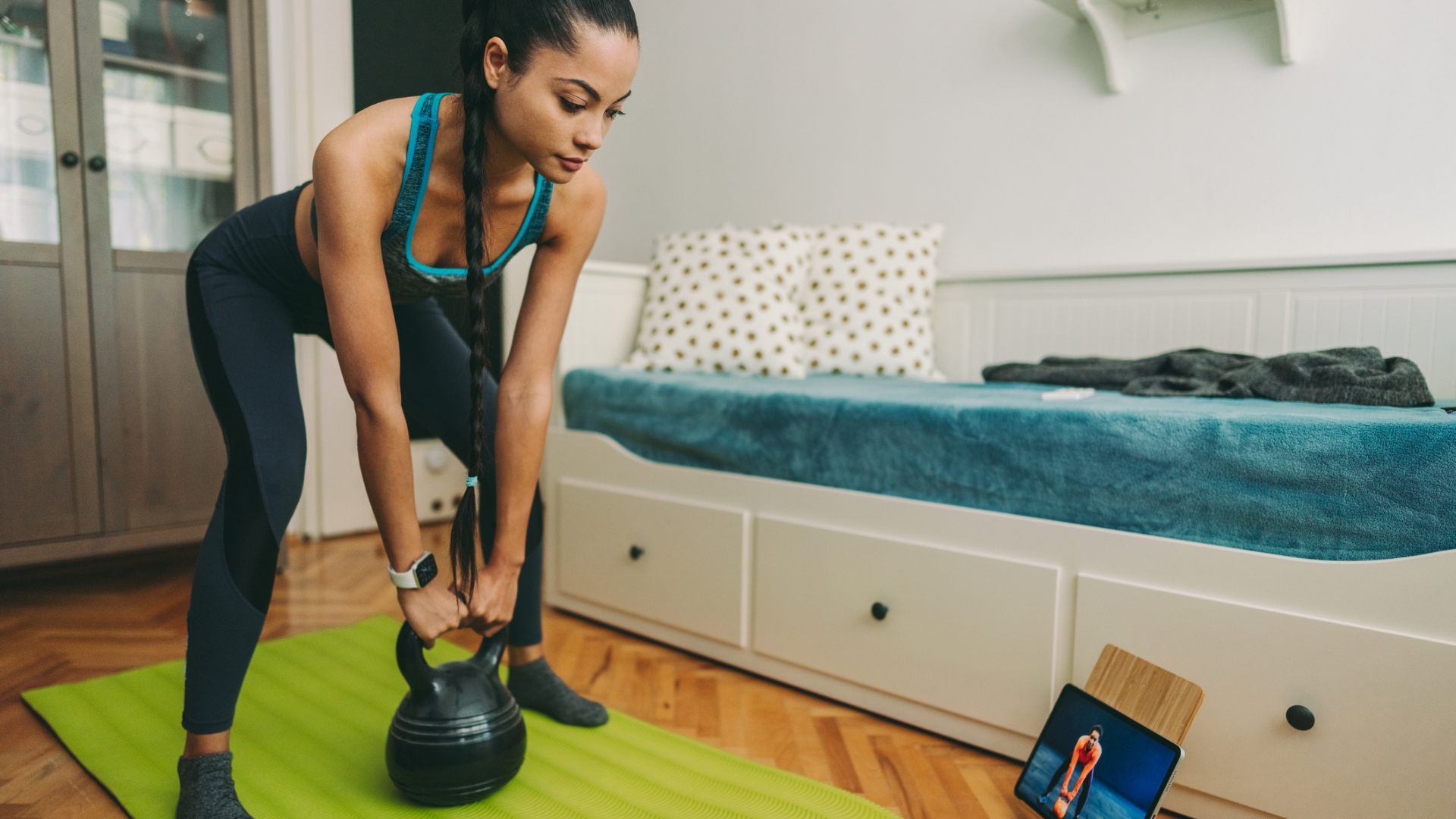 woman standing on a green exercise mat in a home setting holding a kettlebell that's on the floor in between her lets. she's looking at an ipad in front on the floor in front of her.