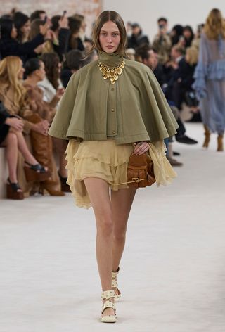 a photo showing the fall jacket trend, capes on the Chloe runway with a model wearing a green cape over a butter yellow ruffle dress styled with butter yellow ballet flats, a gold necklace, and a brown bag