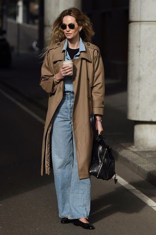 how to make a button-down shirt look more expensive is shown in a photo of a woman standing in the street wearing a tan trench coat over a denim button-down shirt styled with a black t-shirt underneath, baggy jeans, black ballet flats, aviator sunglasses, and a black handbag