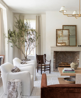 A neutral-toned living room with a large tree, white armchairs and a fireplace.
