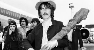 George Harrison pictured on the tarmac beside a plane carrying his sitar in a gigbag. He wears a wide-brimmed hat, white shirt and dark jacket, and sports a moustache.